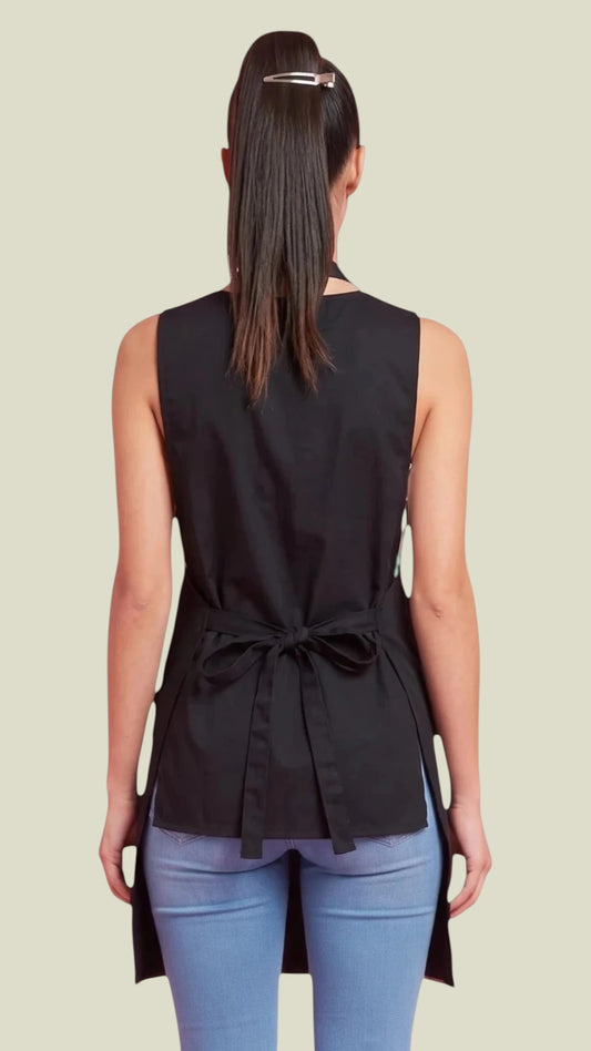 Back view of a dark-haired woman in a ponytail modeling the Ladybird Line 182 Sassy Stylist Rhinestone Apron, showing the tied back waist closure, adjustable halter neck strap, and side waist slots, worn over blue jeans against a solid off-white background.