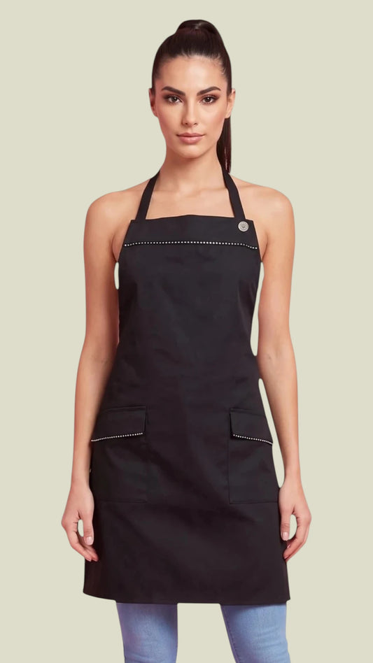 Front view of a dark-haired woman in a ponytail modeling the Ladybird Line 182 Sassy Stylist Rhinestone Apron, featuring a black apron with sparkling rhinestone detailing along the top chest trim and front pockets, worn over blue jeans against a solid off-white background.