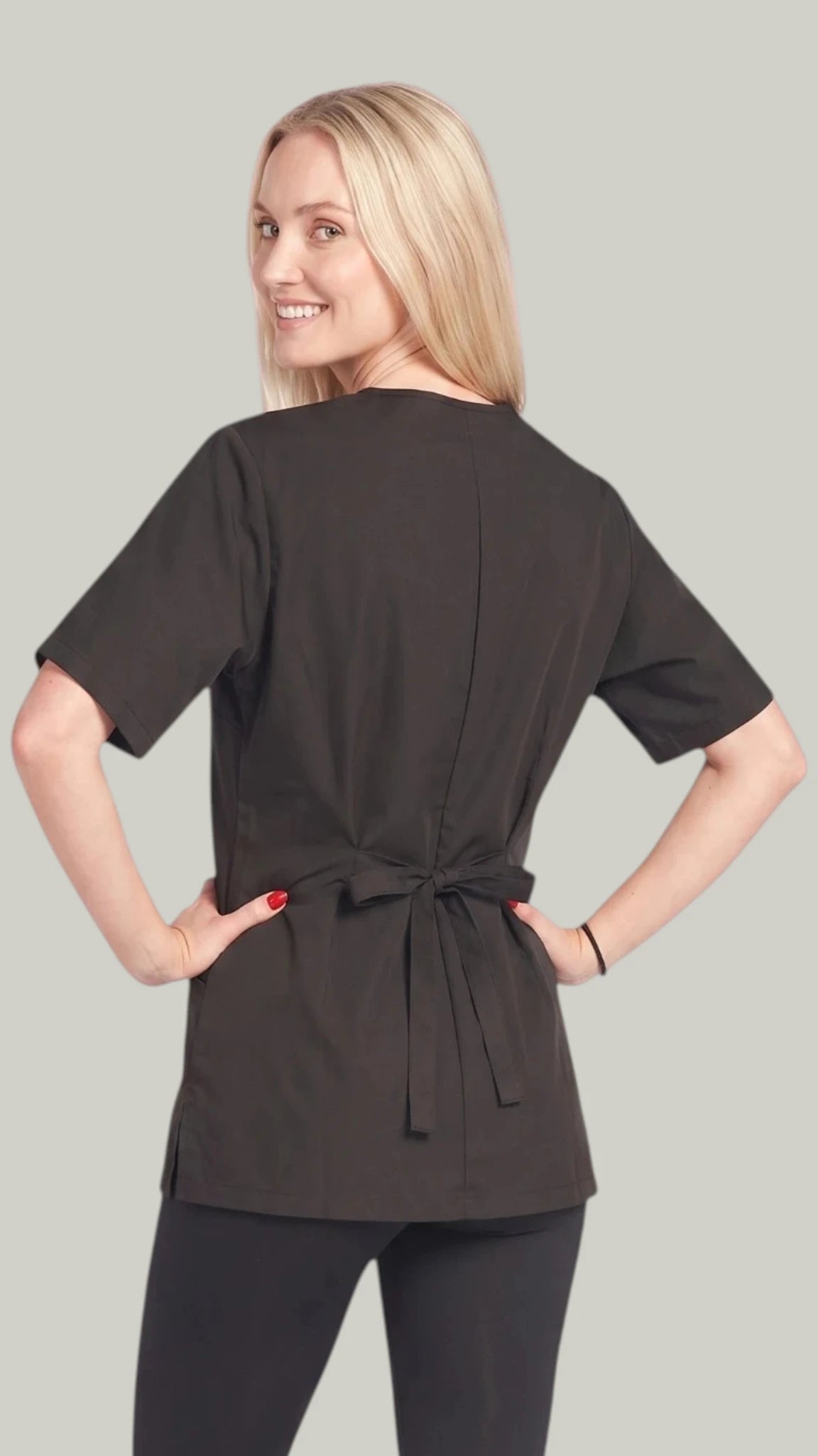 Back view of a blonde woman looking over her shoulder while modeling the Ladybird Line 202 Black Water Resistant Scrub Tunic, featuring a black short-sleeve scrub tunic with an adjustable waist tie belt tied in a bow, worn over black pants against a solid light grey background.