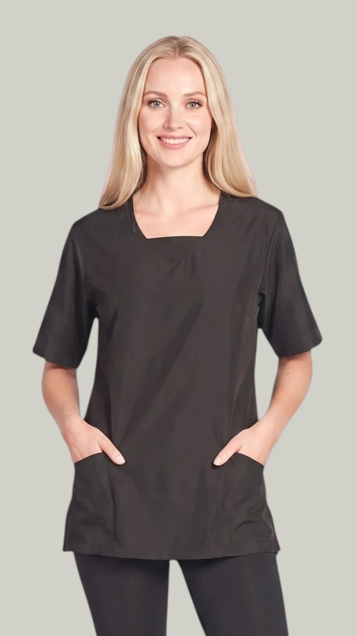 Front view of a smiling blonde woman modeling the Ladybird Line 202 Black Water Resistant Scrub Tunic, featuring a black short-sleeve scrub tunic with a V-neckline and deep front pockets, worn over black pants against a solid light grey background.