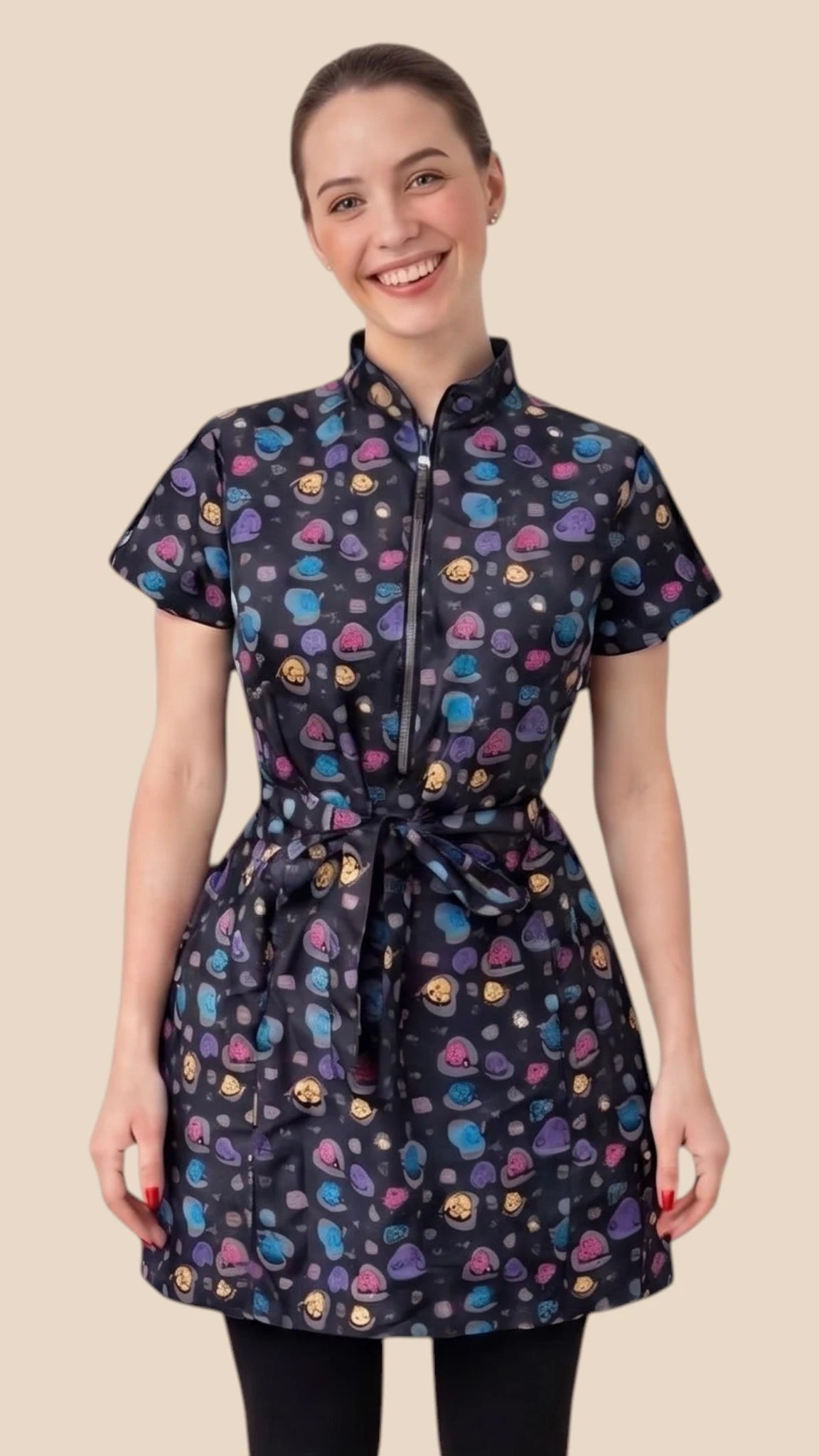 Front view of a dark-haired woman smiling while modeling the Ladybird Line 213 Rainbow Half Zipper Mini Paw Print Water Resistant Scrub Tunic, featuring a black short-sleeve tunic with an all-over colorful mini paw print pattern, rainbow half-zipper, mandarin collar, front pockets, and an adjustable waist tie belt, worn over dark pants against a solid light beige background.