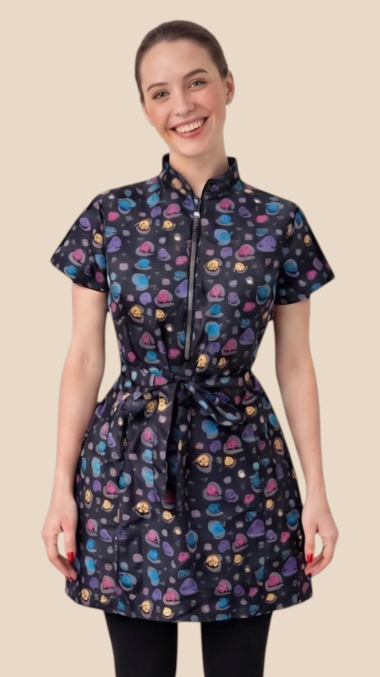 Front view of a dark-haired woman smiling while modeling the Ladybird Line 213 Rainbow Half Zipper Mini Paw Print Water Resistant Scrub Tunic, featuring a black short-sleeve tunic with an all-over colorful mini paw print pattern, rainbow half-zipper, mandarin collar, front pockets, and an adjustable waist tie belt, worn over dark pants against a solid light beige background.