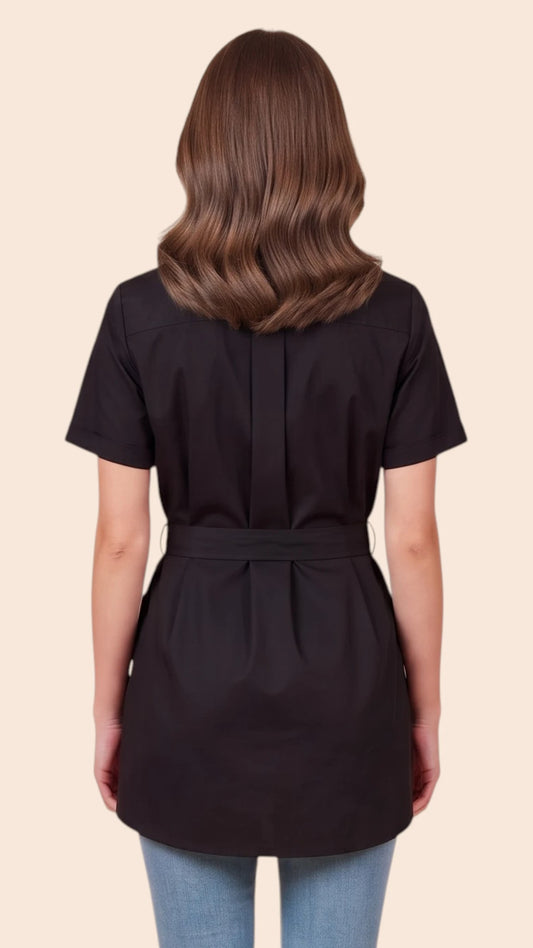 Back view of a brown-haired woman modeling the Ladybird Line 214 Classic Black Rhinestone Half Zipper Water Resistant Scrub Tunic, showing the back tie belt cinched at the waist over light blue jeans against a solid light beige background.