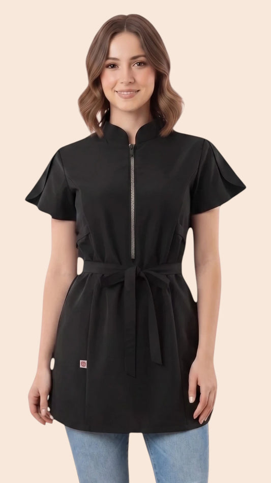 Front view of a brown-haired woman smiling while modeling the Ladybird Line 214 Classic Black Rhinestone Half Zipper Water Resistant Scrub Tunic, featuring a black short-sleeve tunic with a rhinestone half-zipper front, mandarin collar, front pockets, and an adjustable waist tie belt, worn over blue jeans against a solid light beige background.