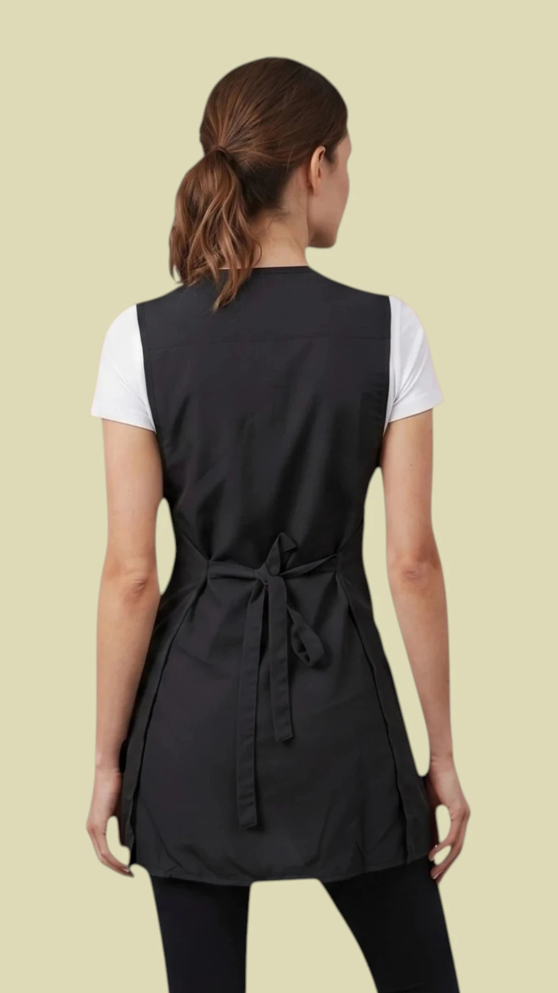 Back view of a woman with her hair in a low ponytail modeling the Ladybird Line 220 Pro Rhinestone Jacket Cobbler Apron, featuring a full-coverage black tunic-length cobbler apron with a back tie belt secured in a bow, worn over a white short-sleeve t-shirt and black pants against a solid light beige background.
