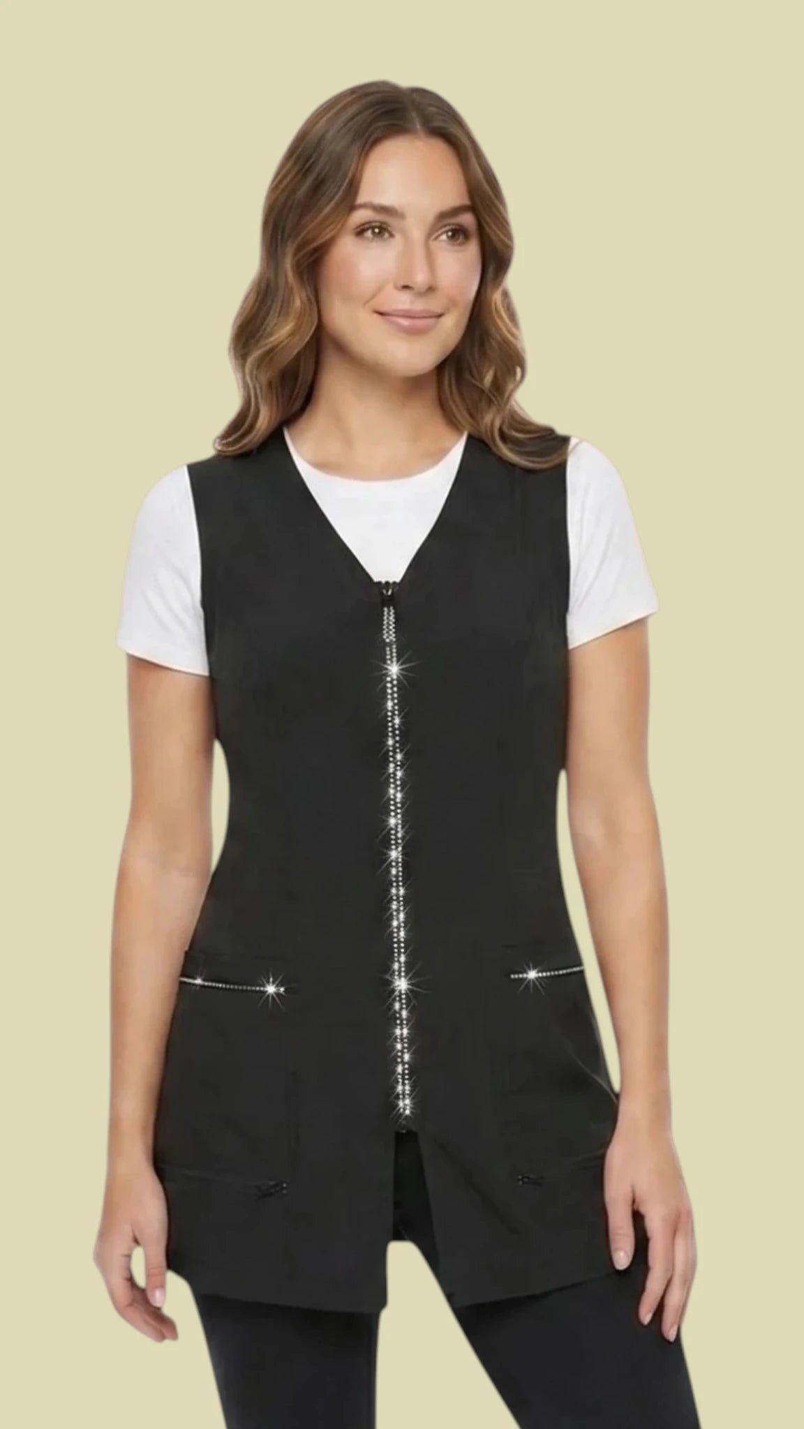 Front view of a smiling woman with wavy light brown hair modeling the Ladybird Line 220 Pro Rhinestone Jacket Cobbler Apron, featuring a black tunic-length cobbler apron with a sparkling rhinestone front zipper, rhinestone-trimmed front pockets, and a V-neckline, worn over a white short-sleeve t-shirt and black pants against a solid light beige background.