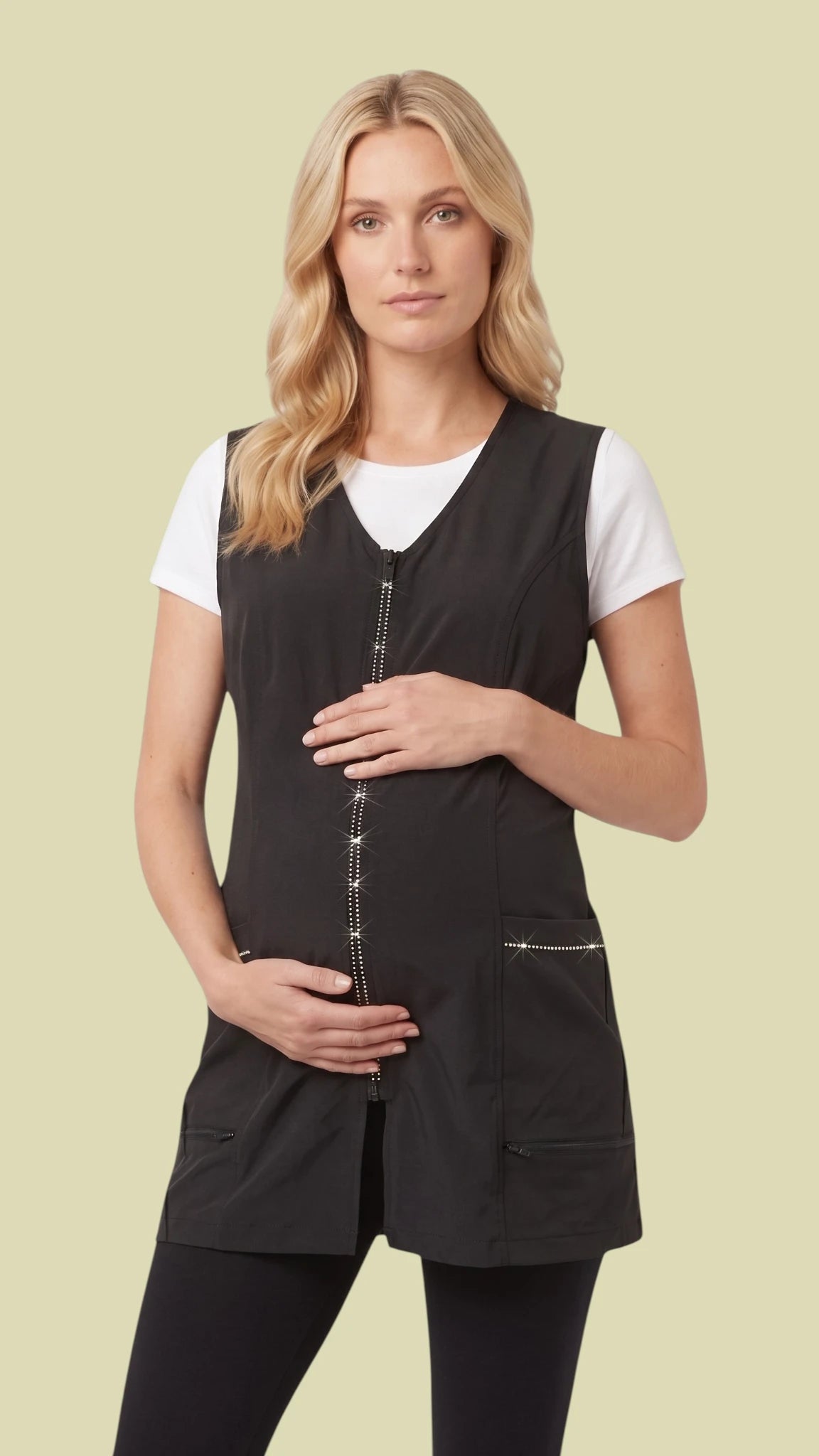 Front view of a pregnant blonde woman modeling the Ladybird Line 220 Pro Rhinestone Jacket Cobbler Apron, featuring a black tunic-length cobbler apron with a sparkling rhinestone front zipper, rhinestone-trimmed front pockets, and a V-neckline, worn over a white short-sleeve t-shirt and black pants against a solid light beige background.