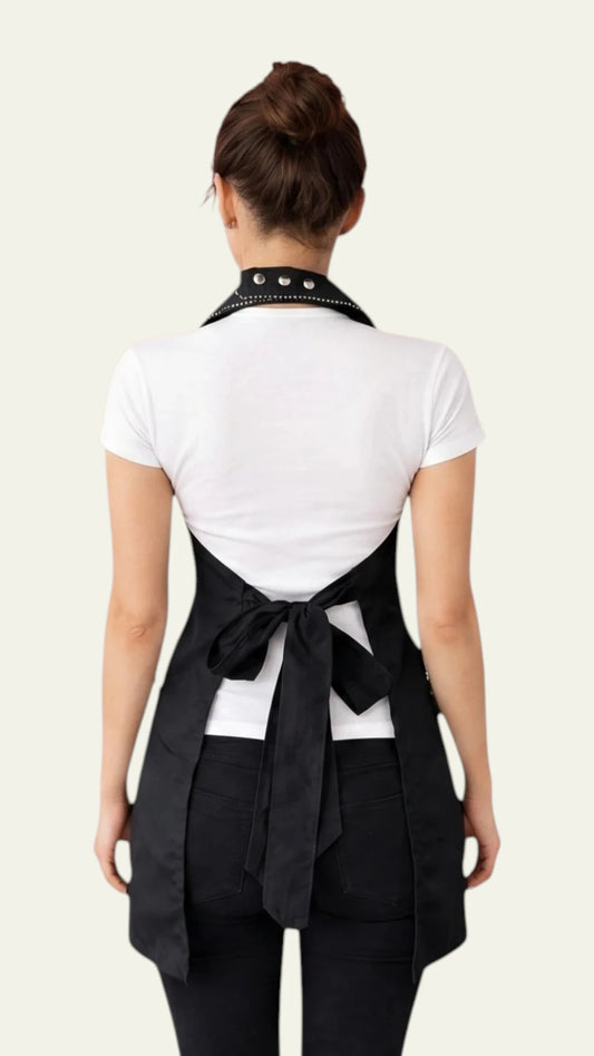 Back view of a woman with her hair in a bun modeling the Ladybird Line 260 Pro Rhinestone Jacket Water Resistant Styling Apron, showing the rhinestone-trimmed collar, open lower back design, and a black waist tie belt secured in a bow, worn over a white short-sleeve t-shirt and black pants against a solid light grey background.