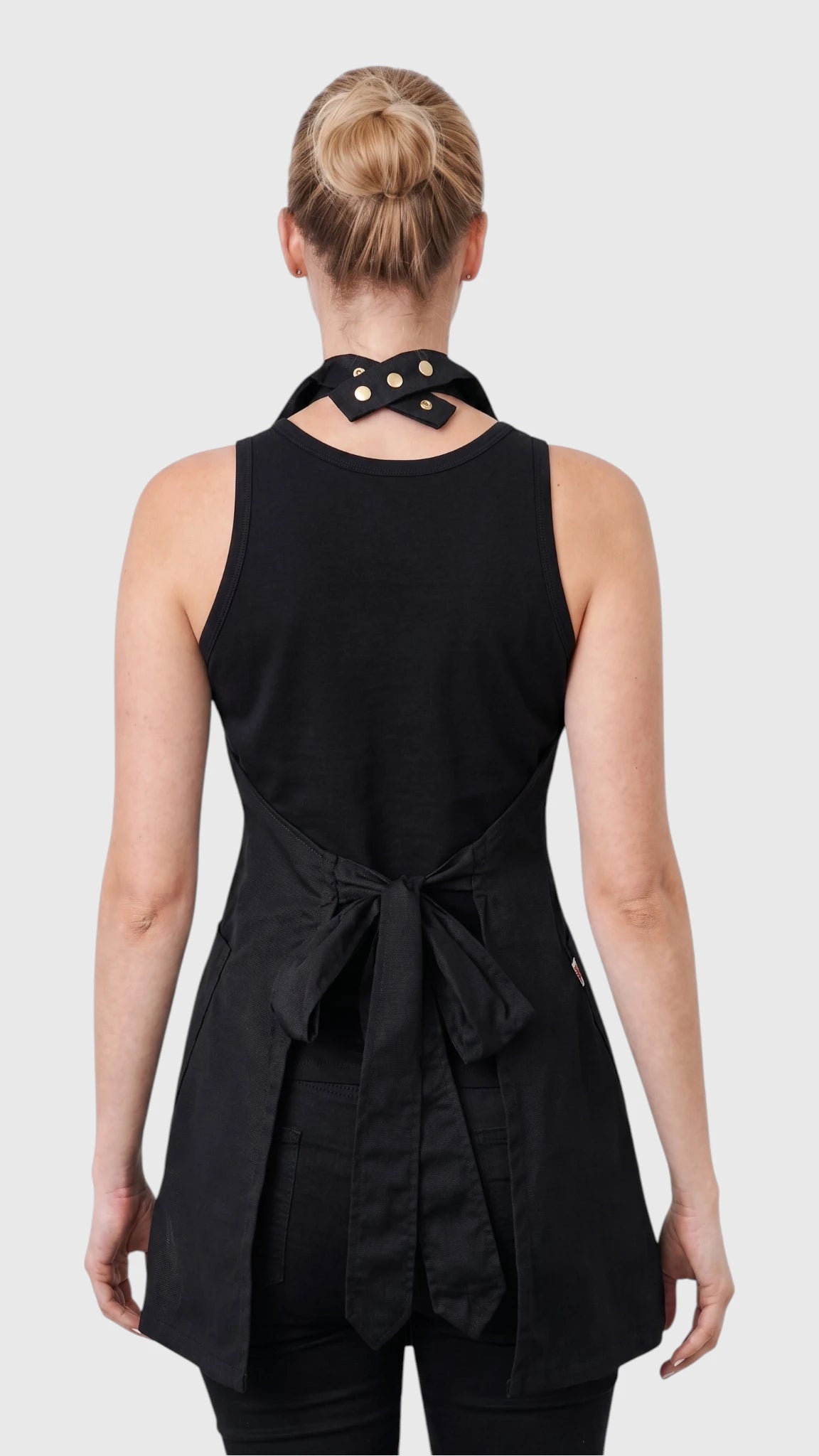Back view of a blonde woman modeling the Ladybird Line 280 Pro Jacket Style Water Resistant Styling Apron, featuring a solid black sleeveless vest-style apron with a high collar, open lower back, and a waist strap tied in a bow, worn over black pants against a solid light grey background.