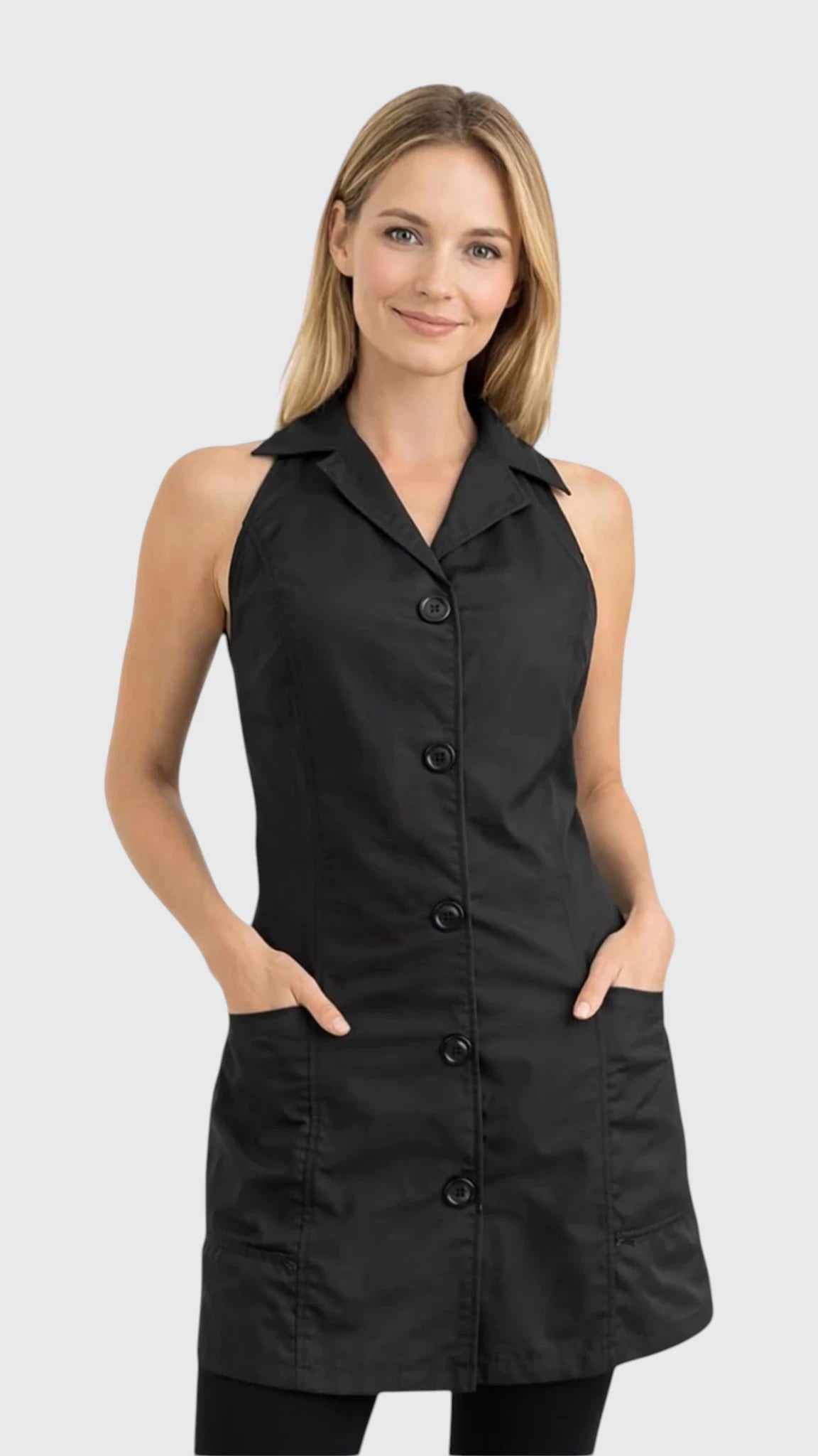 Front view of a smiling blonde woman modeling the Ladybird Line 280 Pro Jacket Style Water Resistant Styling Apron, featuring a black sleeveless vest-style apron with a lapel collar, five decorative front buttons, and deep side pockets, worn over black pants against a solid light grey background.