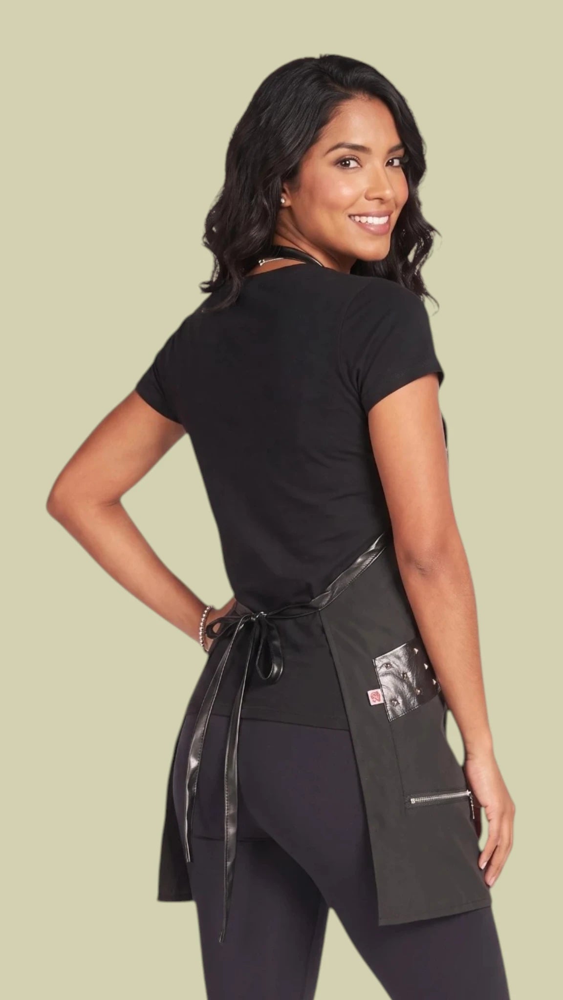 Back three-quarter view of a smiling woman with long dark hair modeling the Ladybird Line 315 Metal Stud Water Resistant Styling Apron, showing the back crisscross adjustable neck strap, waist tie belt tied in a bow, faux-leather trim along the pockets, and side zippered pocket, worn over a black t-shirt and black pants against a solid light beige background.