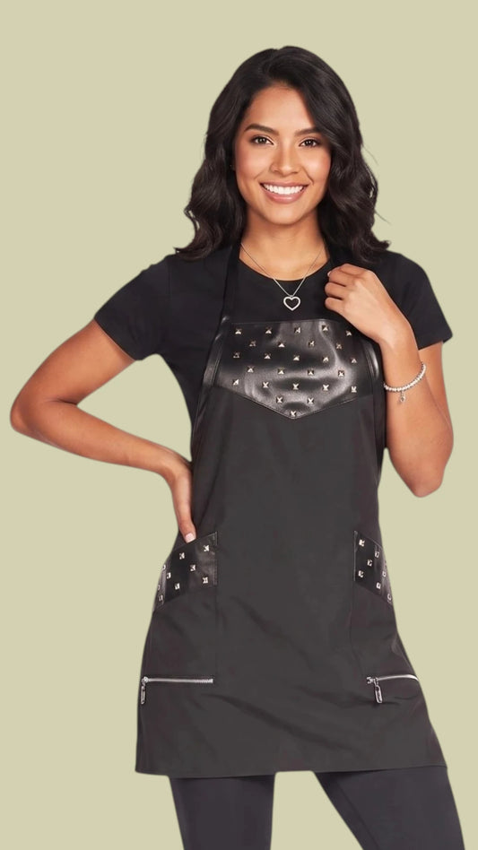Front view of a smiling woman with long dark hair modeling the Ladybird Line 315 Metal Stud Water Resistant Styling Apron, featuring a black apron with faux-leather trim and square metal studs on the chest and upper pockets, deep zippered front pockets, and an adjustable neck strap, worn over a black t-shirt and black pants against a solid light beige background.