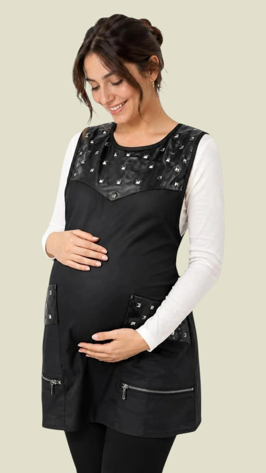 Front view of a smiling woman modeling the Ladybird Line 345 Metal Stud Water Resistant Cobbler Apron, featuring a black cobbler apron with faux-leather trim and square silver studs on the upper chest and pockets, deep zippered pockets, and side ties, layered over a white long-sleeve shirt and black leggings against a solid light beige background.