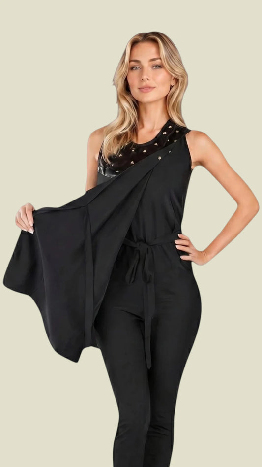 Front view of a blonde woman posing in the Ladybird Line 345 Metal Stud Water Resistant Cobbler Apron, holding out the front panel drape to show the full coverage, featuring black fabric with faux-leather trim and square metal studs on the upper chest, layered over a black jumpsuit against a solid light beige background.