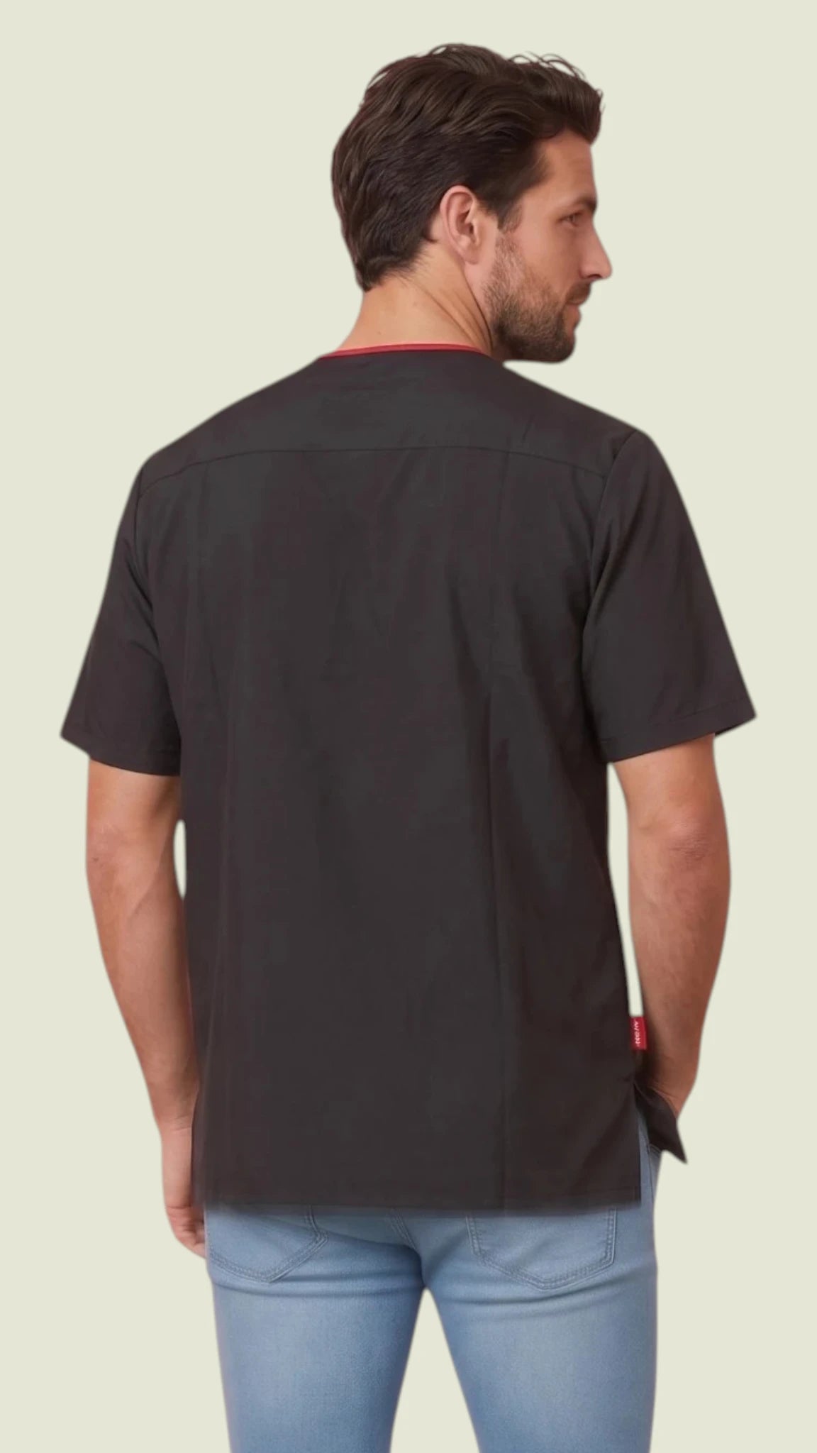 Back view of a man with short brown hair and a beard modeling the Ladybird Line 415 Men's Waterproof & Bleach Resistant Short Sleeve Scrub Top, showing the solid black back panel and red inner collar accent, worn over light blue jeans against a light beige background.