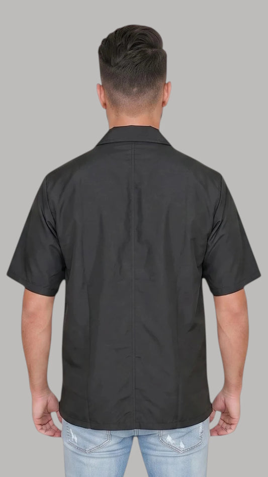 Back view of a man with short dark hair modeling the Ladybird Line 415 Unisex Barber & Grooming Water Resistant Jacket, showing the solid black short-sleeve jacket back with a crisp collar, worn over light blue jeans against a solid light grey background.