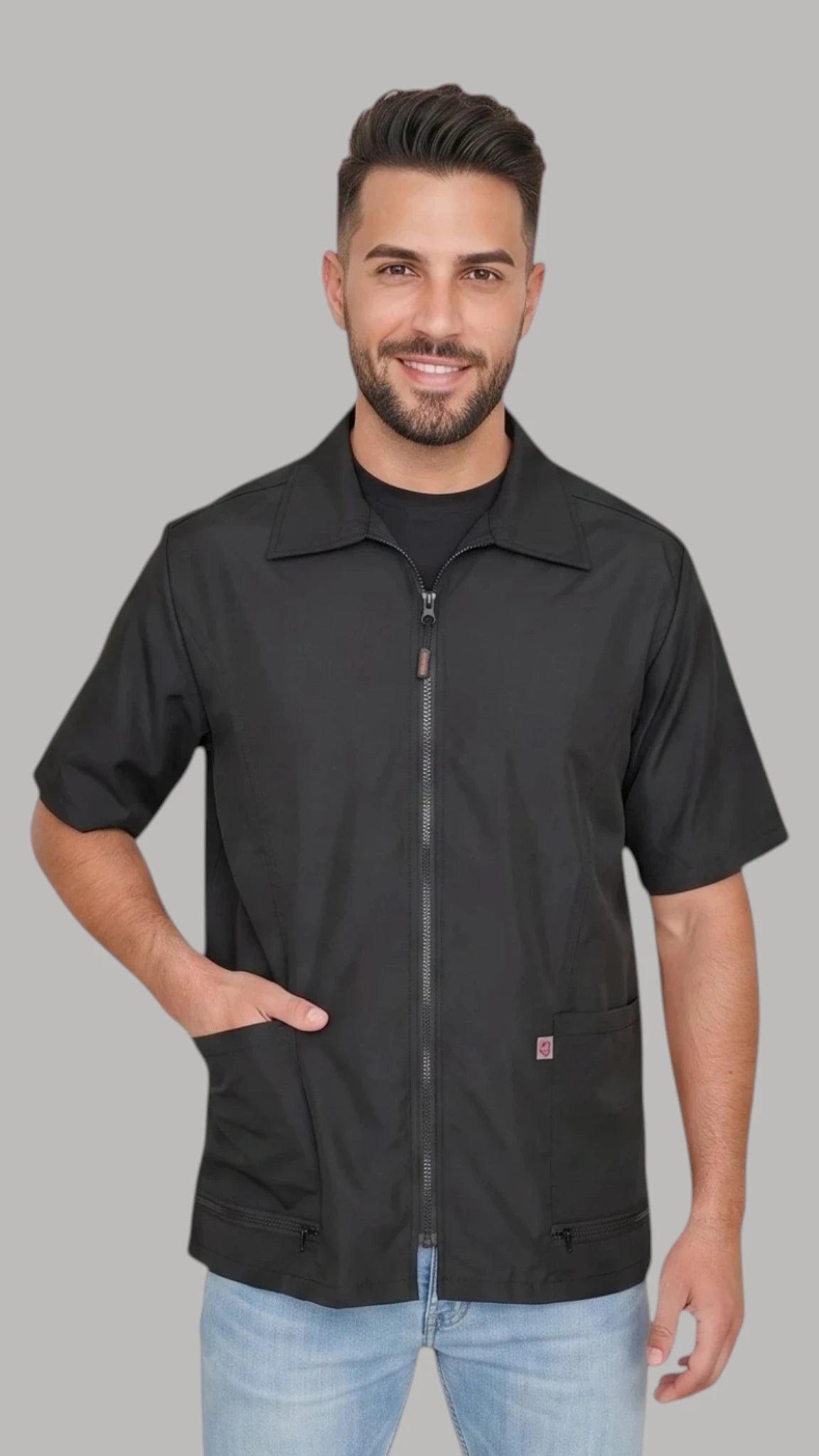 Front view of a smiling man with short dark hair and a beard modeling the Ladybird Line 415 Unisex Barber & Grooming Water Resistant Jacket, featuring a black short-sleeve collared jacket with a full front zipper closure, deep side pockets with zippered bottoms, and a brand logo tag, worn over light blue jeans against a solid light grey background.