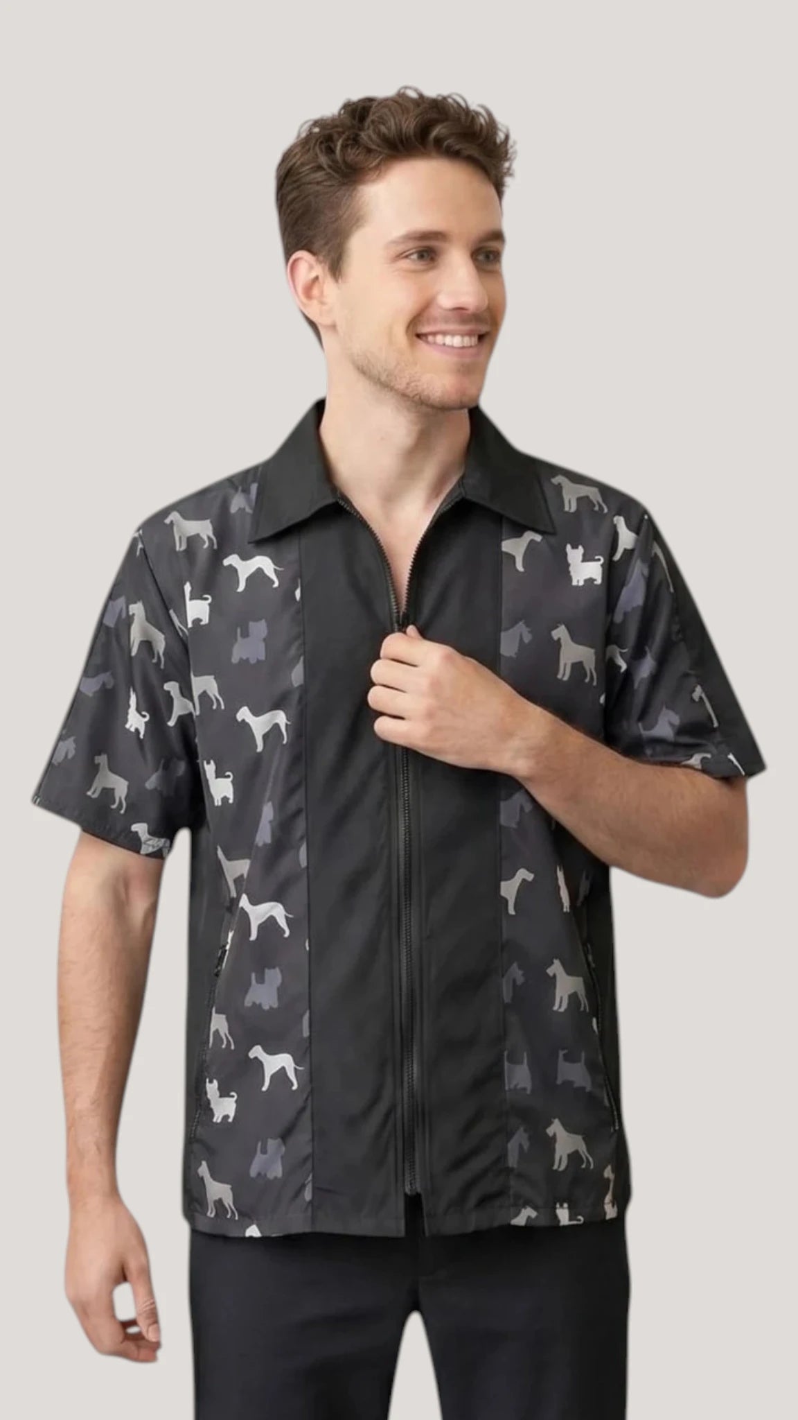 Front view of a brown-haired man smiling and modeling the Ladybird Line 420 Men's Terrier Print Water Resistant Grooming Jacket, featuring a black short-sleeve jacket with a shirt collar, front zipper closure, deep side pockets, and charcoal side panels printed with grey dog breed silhouettes, worn over black pants against a solid light grey background.