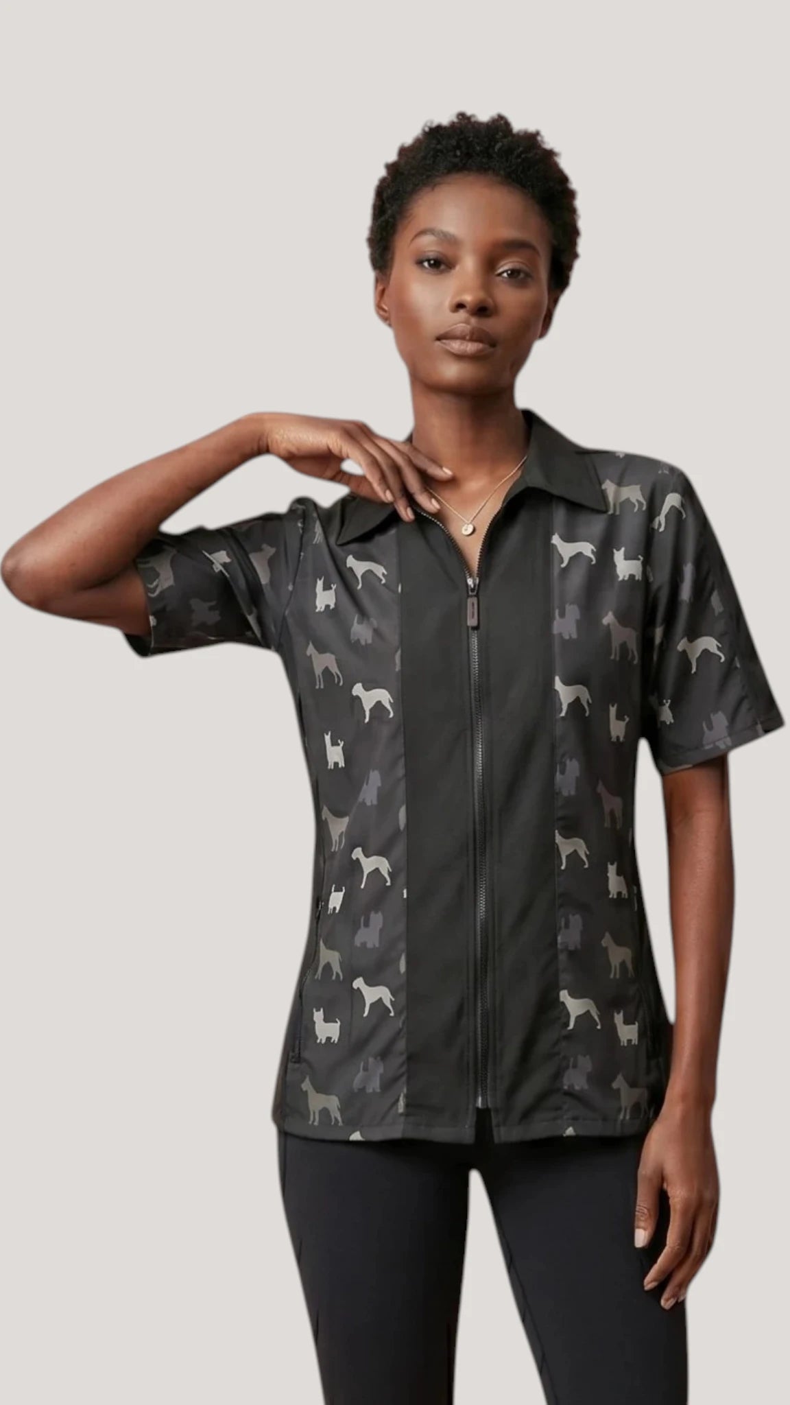 Front view of a dark-haired woman modeling the Ladybird Line 420 Men's Terrier Print Water Resistant Grooming Jacket, featuring a black short-sleeve collared jacket with a front zipper closure and dark charcoal panels printed with light grey terrier dog breed silhouettes, worn over black leggings against a solid light grey background.