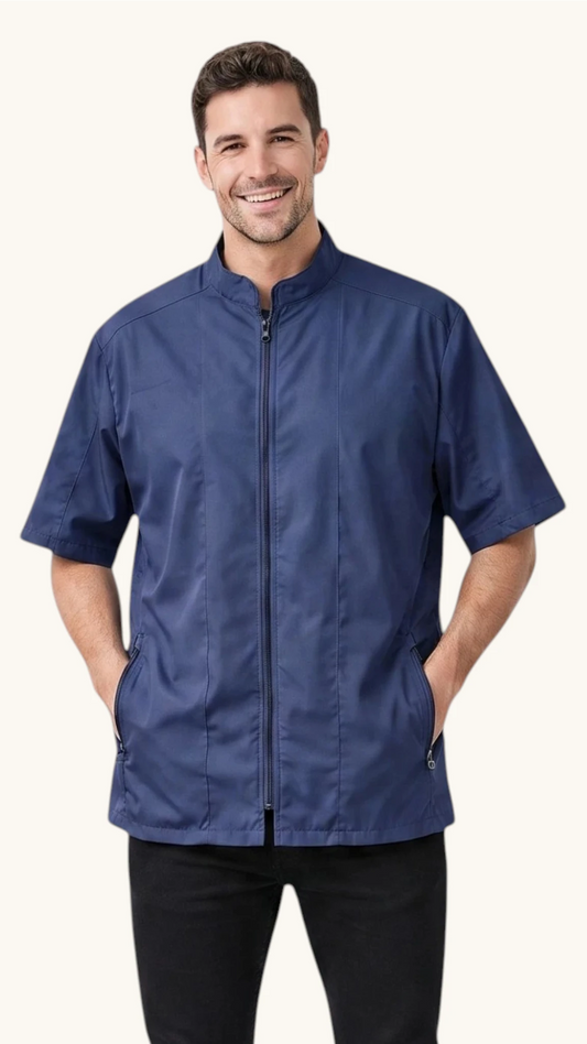 Front view of a dark-haired man smiling with his hands in his pockets while modeling the Ladybird Line 427 Water Resistant Navy Mandarin Collar Tailored Barber & Grooming Jacket, featuring a navy zip-up short-sleeve jacket with a mandarin collar, paired with black pants against a solid off-white background.