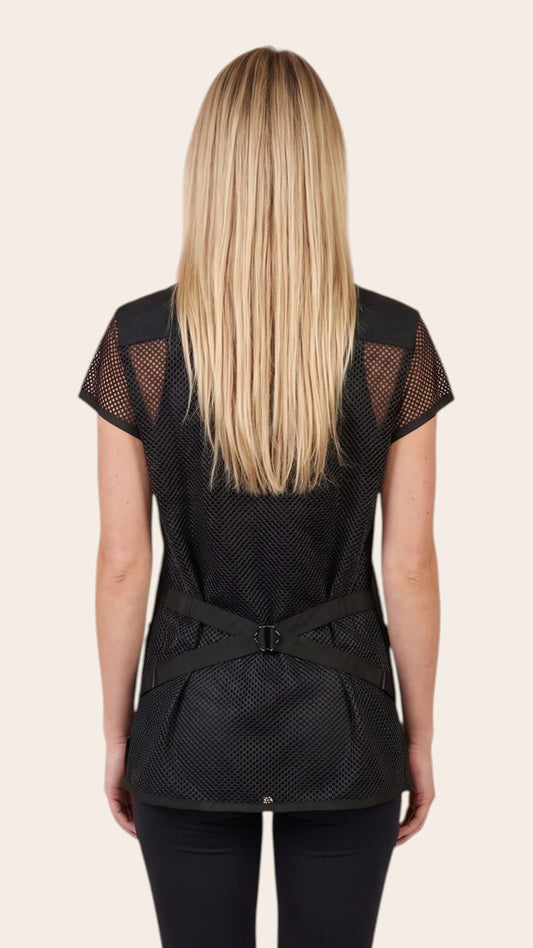 Back view of a blonde woman modeling the Ladybird Line 430 Women's Mesh Back Water & Hair Resistant Snap Styling Jacket, showing the black jacket back featuring a full mesh panel, mesh short sleeves, and an adjustable back tie belt, worn over black pants against a solid light beige background.