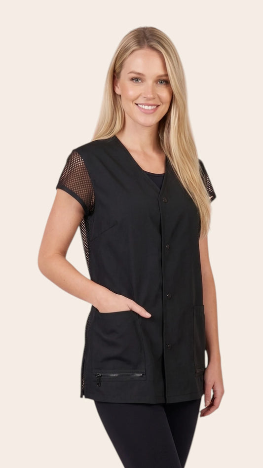 Front view of a smiling blonde woman modeling the Ladybird Line 430 Women's Mesh Back Water & Hair Resistant Snap Styling Jacket, featuring a black short-sleeve jacket with mesh shoulder sleeves, snap-button closure, and deep front patch pockets, worn over black pants against a solid light beige background.