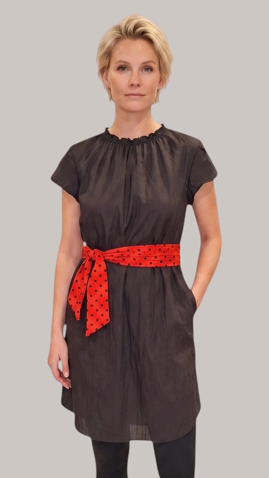 Front view of a blonde woman modeling the Ladybird Line 444 Classic Black Water Resistant Scarf Belt Professional Dress, featuring a black short-sleeve dress with a gathered neckline, elastic waist, side pockets, and a red polka dot scarf belt, worn over dark pants against a solid light grey background.
