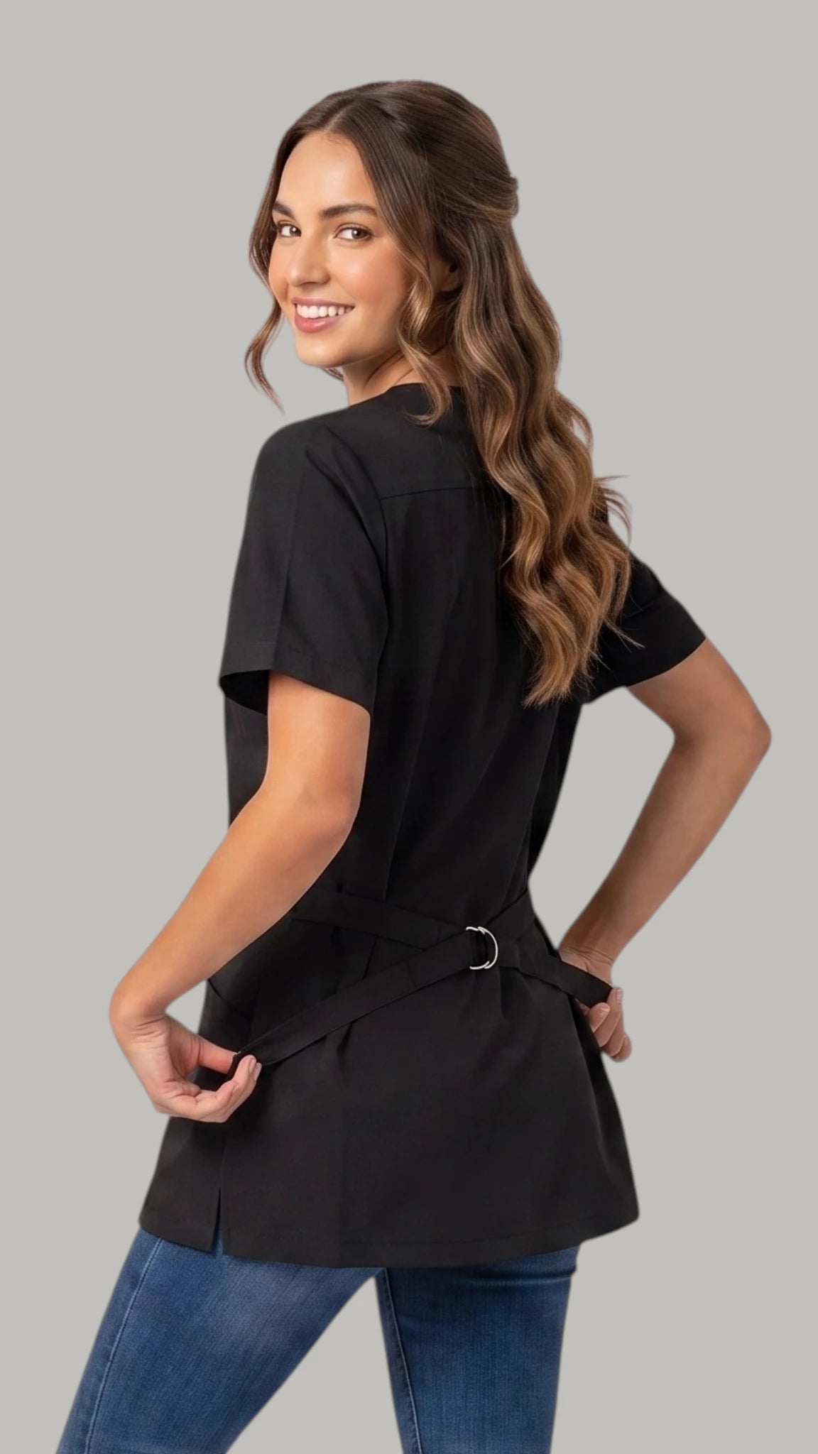 Back view of a brown-haired woman looking back over her shoulder while modeling the Ladybird Line 460 Women's Rhinestone Zipper Water & Hair Resistant Grooming Jacket, showing the black short-sleeve jacket back with an adjustable back belt tied at the waist, worn over blue jeans against a solid light grey background.