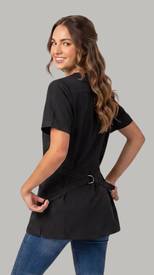 Back view of a brown-haired woman looking back over her shoulder while modeling the Ladybird Line 460 Women's Rhinestone Zipper Water & Hair Resistant Grooming Jacket, showing the black short-sleeve jacket back with an adjustable back belt tied at the waist, worn over blue jeans against a solid light grey background.