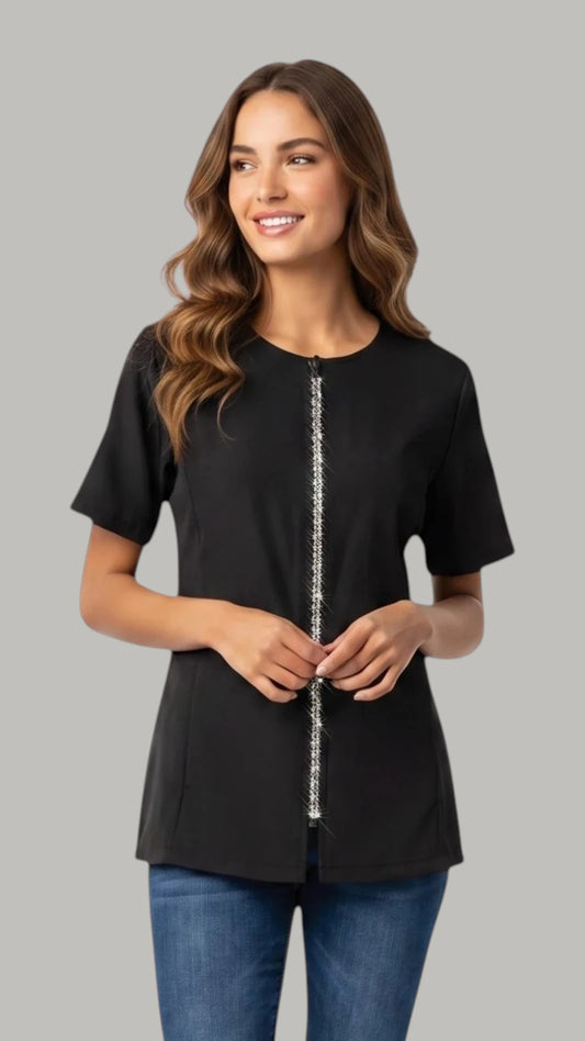 Front view of a brown-haired woman smiling and modeling the Ladybird Line 460 Women's Rhinestone Zipper Water & Hair Resistant Grooming Jacket, featuring a black short-sleeve jacket with a round collar, sparkling rhinestone zipper closure, and deep side pockets, worn over blue jeans against a solid light grey background.