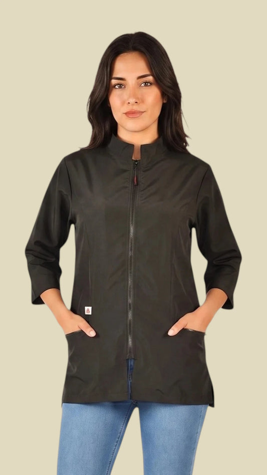 Front view of a dark-haired woman posing with hands in pockets modeling the Ladybird Line 512 Women's Black 3/4 Sleeve Water Resistant Stylist Jacket, featuring a black jacket with a mandarin collar, front zip closure, 3/4 sleeves, and deep side pockets, worn over blue jeans against a solid light beige background.