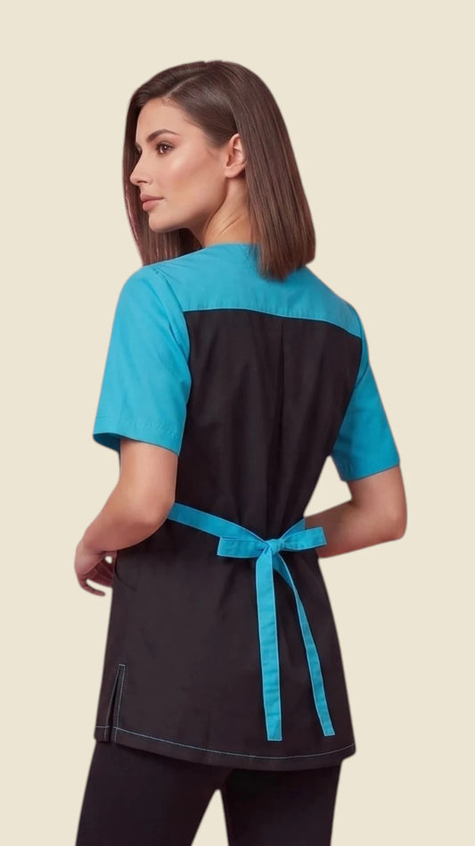 Back view of a dark-haired woman looking back over her shoulder while modeling the Ladybird Line 515 Women's Blue Rhinestone Zipper Color Block Styling Jacket, showing the black jacket back with bright turquoise blue short sleeves, upper shoulder yoke, and tied back waist sash, worn over black pants against a solid light beige background.