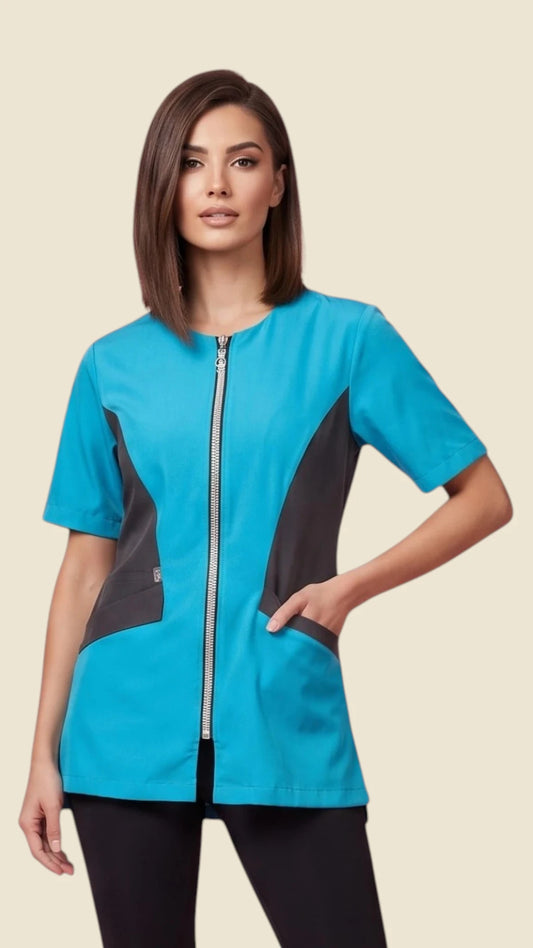 Front view of a dark-haired woman modeling the Ladybird Line 515 Women's Blue Rhinestone Zipper Color Block Styling Jacket, featuring a bright turquoise blue short-sleeve smock with black color-blocked side panels, rhinestone front zipper closure, and deep side pockets, worn over black pants against a solid light beige background.