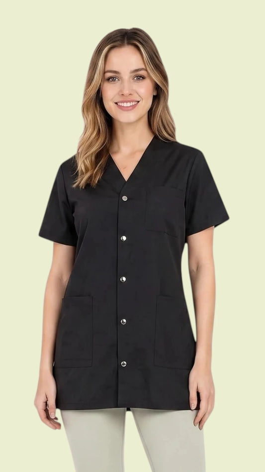 Front view of a smiling woman with blonde hair modeling the Ladybird Line 530 Pro Salon Water Resistant & Bleach Resistant Styling Jacket, featuring a black short-sleeve jacket with a V-neck, snap-button closure, and two deep front patch pockets, worn over light grey pants against a solid pale yellow background.