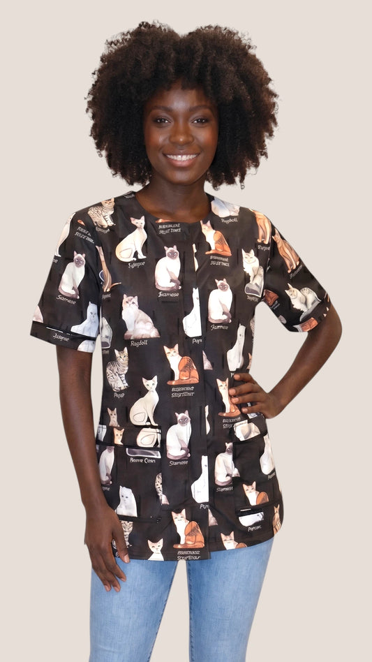 Front view of a dark-skinned woman with curly hair smiling while modeling the Ladybird Line 545 The Catty Groomer Waterproof & Bleach Resistant Grooming Jacket, featuring a black short-sleeve grooming smock with an all-over colorful cat breed print, worn over blue jeans against a solid off-white background.