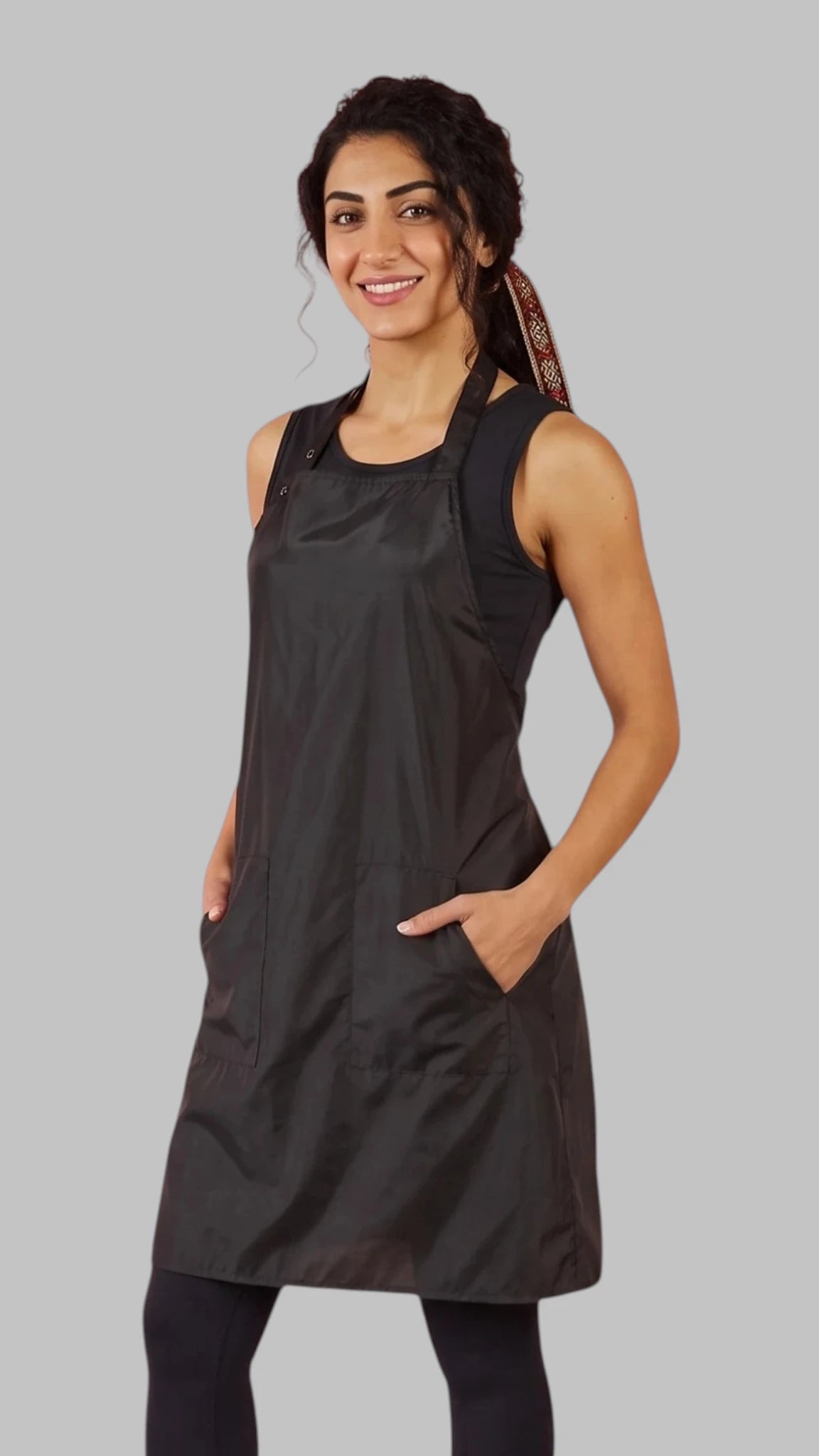 Front view of a dark-haired woman smiling with hands in pockets modeling the Ladybird Line 600 Women's Classic Black Waterproof & Hair Resistant Bather's Grooming Apron, featuring a solid black apron with an adjustable neck strap and deep side pockets, worn over a black tank top and black leggings against a light grey background.