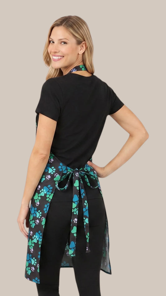Back view of a blonde woman looking back over her shoulder while modeling the Ladybird Line 607 Unisex Blue Paw Print Waterproof & Hair Resistant Bather's Grooming Apron, showing the back tie belt tied into a bow at the waist over a black t-shirt and black pants against a solid light beige background.