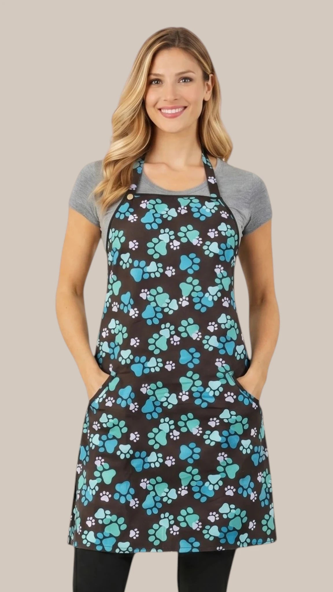Front view of a smiling blonde woman posing with hands in pockets modeling the Ladybird Line 607 Unisex Blue Paw Print Waterproof & Hair Resistant Bather's Grooming Apron, featuring a black apron with a repeating pattern of blue, teal, and white paw prints, adjustable neck band, and deep side pockets, worn over a grey t-shirt and black pants against a solid light beige background.