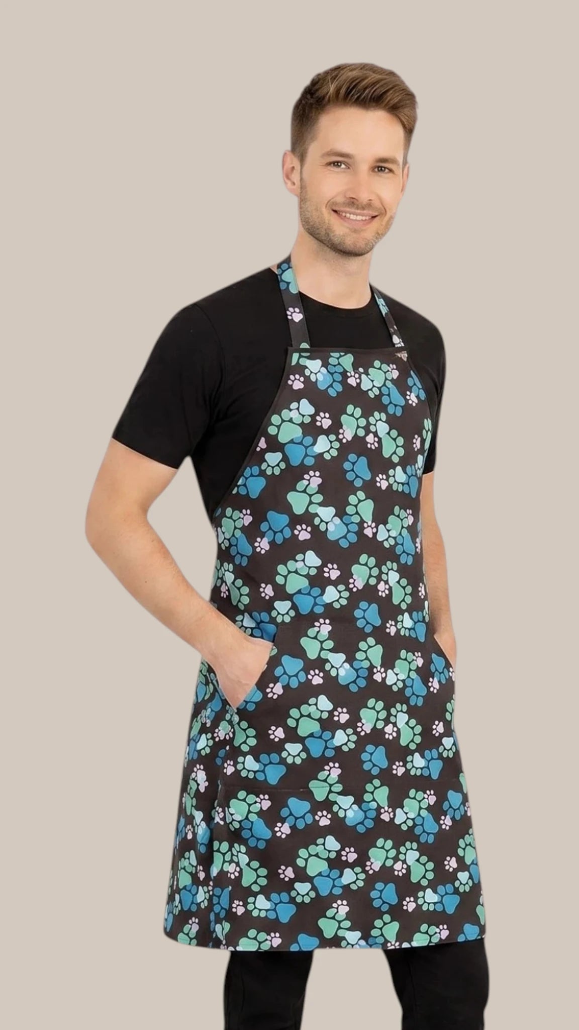 Front view of a dark-haired man posing with hands in pockets modeling the Ladybird Line 607 Unisex Blue Paw Print Waterproof & Hair Resistant Bather's Grooming Apron, featuring a black apron with a repeating pattern of blue, teal, and white paw prints, adjustable neck band, and deep side pockets, worn over a black t-shirt and black pants against a solid light beige background.