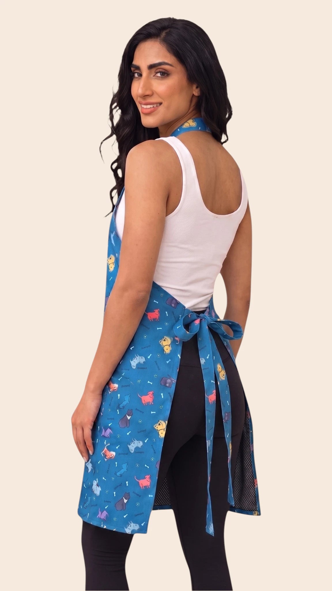 Back view of a dark-haired woman looking back over her shoulder while modeling the Ladybird Line 608 Blue Dog Design Waterproof & Hair Resistant Bather's Grooming Apron, showing the back tie belt tied into a bow at the waist over a white tank top and black leggings against a solid light beige background.