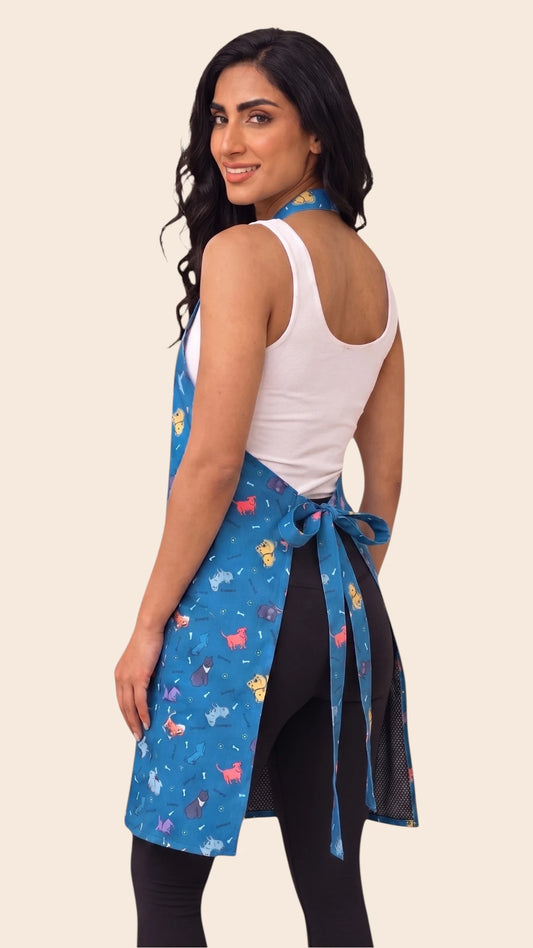 Back view of a dark-haired woman looking back over her shoulder while modeling the Ladybird Line 608 Blue Dog Design Waterproof & Hair Resistant Bather's Grooming Apron, showing the back tie belt tied into a bow at the waist over a white tank top and black leggings against a solid light beige background.