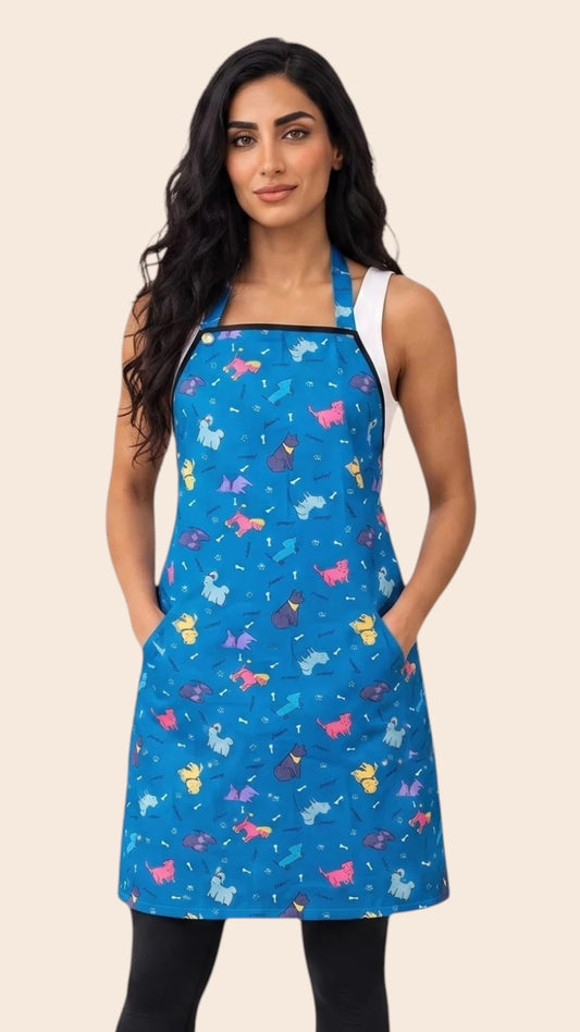 Front view of a dark-haired woman posing with hands in pockets modeling the Ladybird Line 608 Blue Dog Design Waterproof & Hair Resistant Bather's Grooming Apron, featuring a bright blue apron with a colorful repeating pattern of dog silhouettes, adjustable neck strap, and deep side pockets, worn over a white tank top and black pants against a solid light beige background.