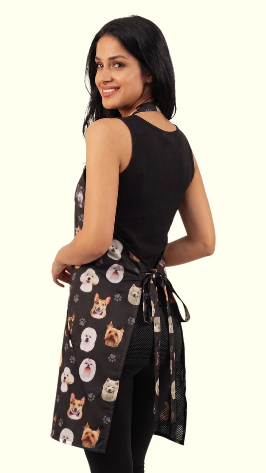 Back view of a dark-haired woman modeling the Ladybird Line 610 Dog Breeds Print Waterproof Hair & Bleach Resistant Grooming Apron, showing the back tie closure, mesh lining, and side towel loop, worn with a black sleeveless top and black pants against a solid off-white background.