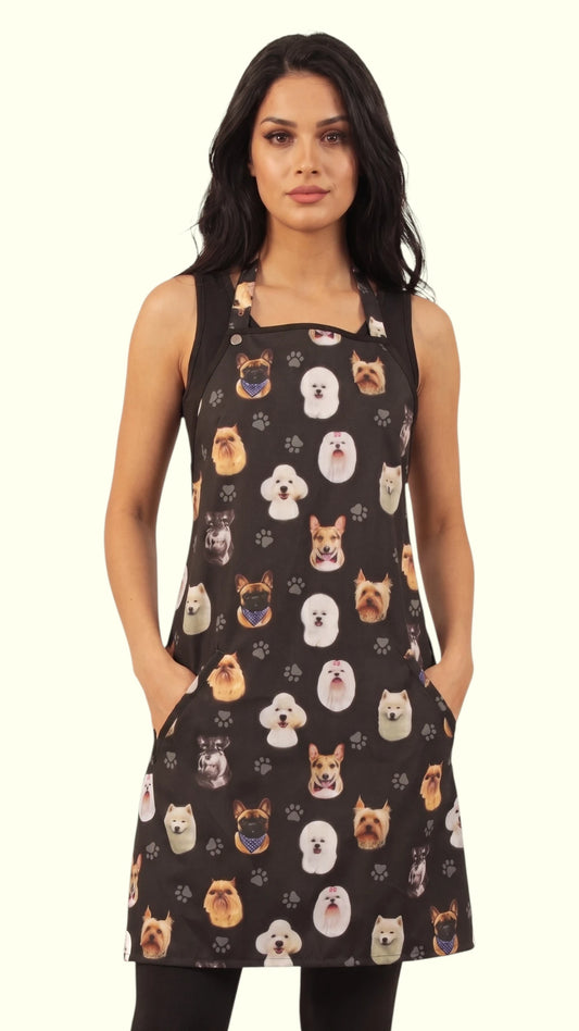 Front view of a dark-haired woman modeling the Ladybird Line 610 Dog Breeds Print Waterproof Hair & Bleach Resistant Grooming Apron, featuring a black apron with an all-over colorful dog breed head pattern, front pockets, and adjustable straps, set against a solid off-white background.