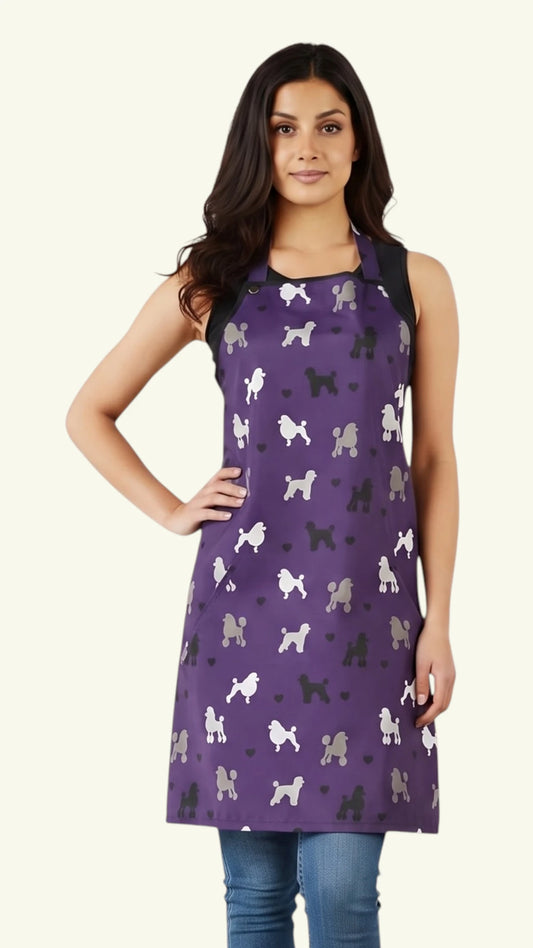 Front view of a dark-haired woman posing with hand on hip modeling the Ladybird Line 611 Purple Poodle Print Waterproof & Bleach Resistant Grooming Apron, featuring a purple apron with a repeating pattern of white, black, and grey poodle silhouettes, adjustable neck strap, and deep side pockets, worn over a black tank top and blue jeans against a light beige background.
