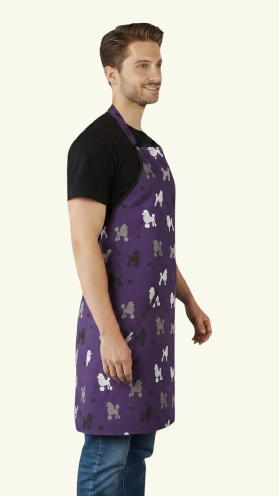 Side profile view of a dark-haired man modeling the Ladybird Line 611 Unisex Purple Poodle Print Waterproof & Bleach Resistant Grooming Apron, featuring a purple apron with a repeating pattern of white, black, and grey poodle silhouettes, worn over a black t-shirt and blue jeans against a light beige background.