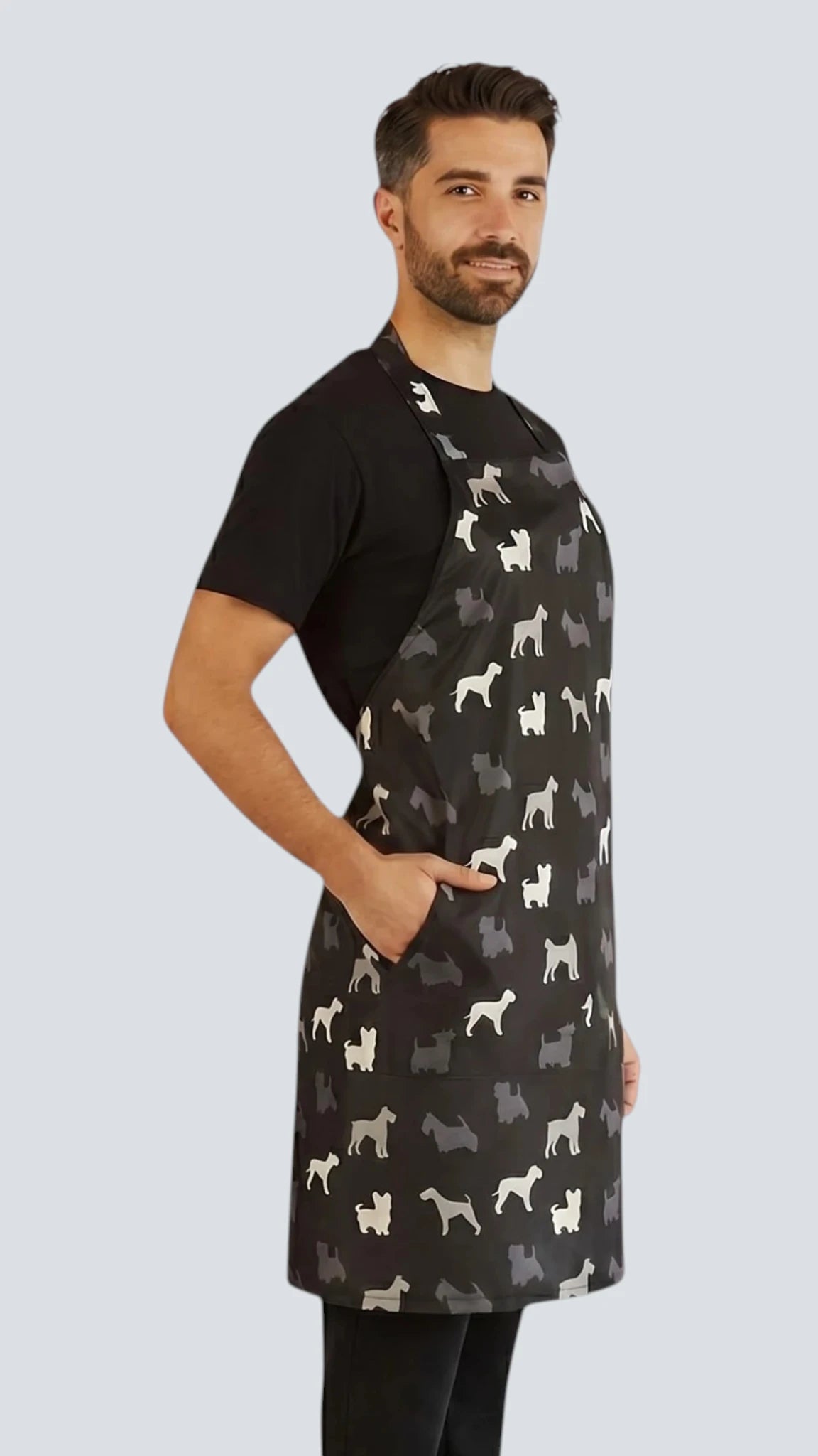 Three-quarter side view of a dark-haired man posing with hand in pocket modeling the Ladybird Line 613 Unisex Terrier Breed Print Waterproof & Bleach Resistant Grooming Apron, featuring a black apron with a repeating white and grey terrier silhouette print, worn over a black t-shirt and black pants against a light grey background.