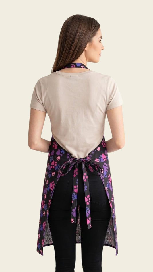 Back view of a brown-haired woman modeling the Ladybird Line 614 Pink & Purple Paw Print Waterproof Grooming Apron, showing the adjustable neck strap and back tie belt tied around the waist over a beige t-shirt and black pants against a solid light beige background.