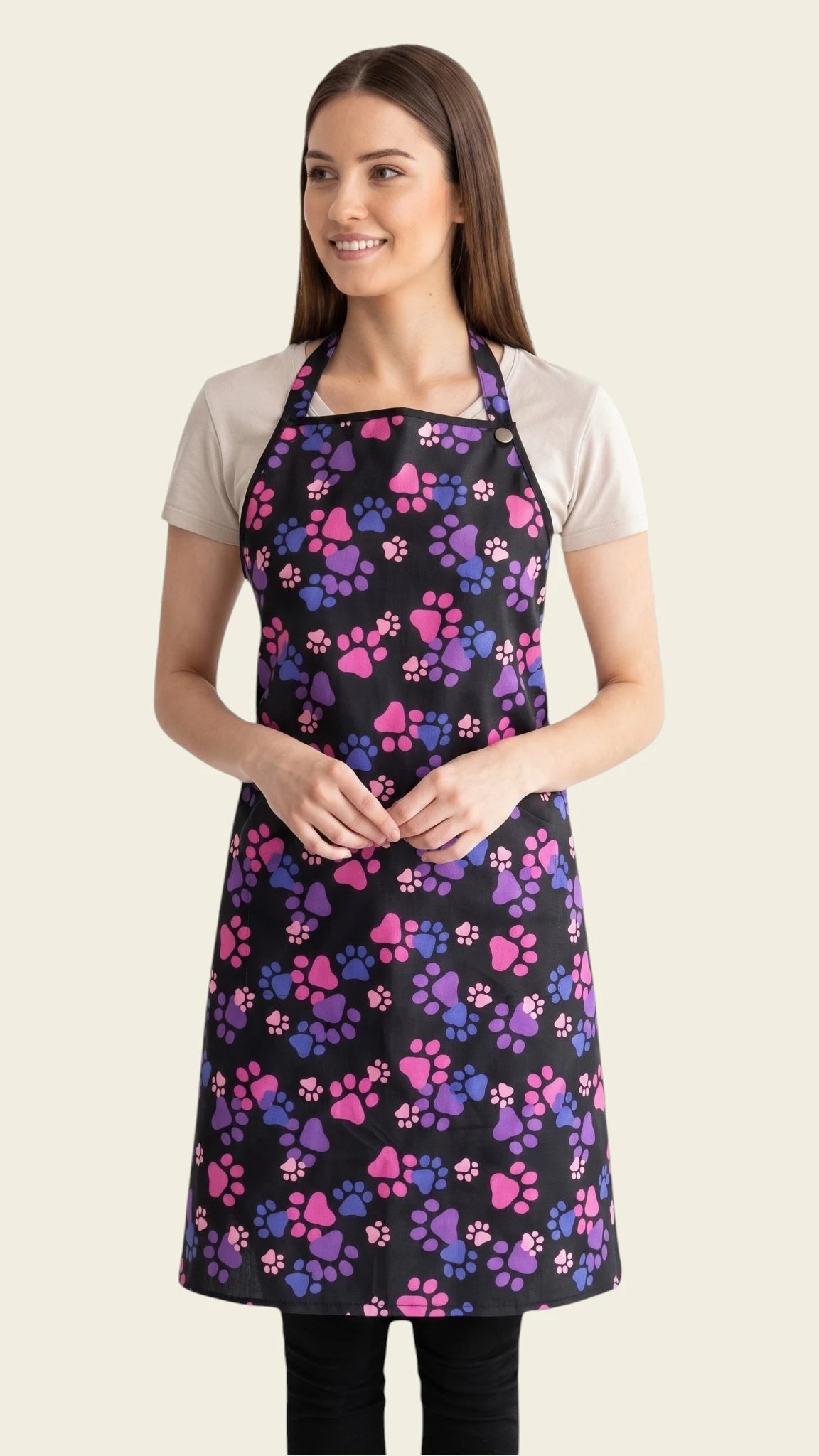 Front view of a brown-haired woman smiling and modeling the Ladybird Line 614 Pink & Purple Paw Print Waterproof Grooming Apron, featuring a full-coverage black apron with a vibrant pink, purple, and blue paw print pattern, adjustable neck strap, back tie belt, and deep side pockets, worn over a beige t-shirt and black pants against a solid light beige background.