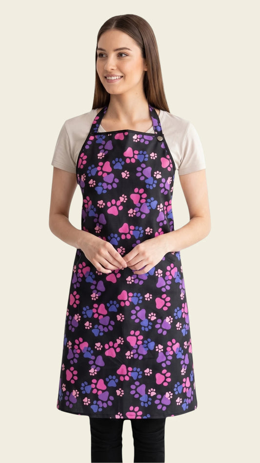 Front view of a brown-haired woman smiling and modeling the Ladybird Line 614 Pink & Purple Paw Print Waterproof Grooming Apron, featuring a full-coverage black apron with a vibrant pink, purple, and blue paw print pattern, adjustable neck strap, back tie belt, and deep side pockets, worn over a beige t-shirt and black pants against a solid light beige background.